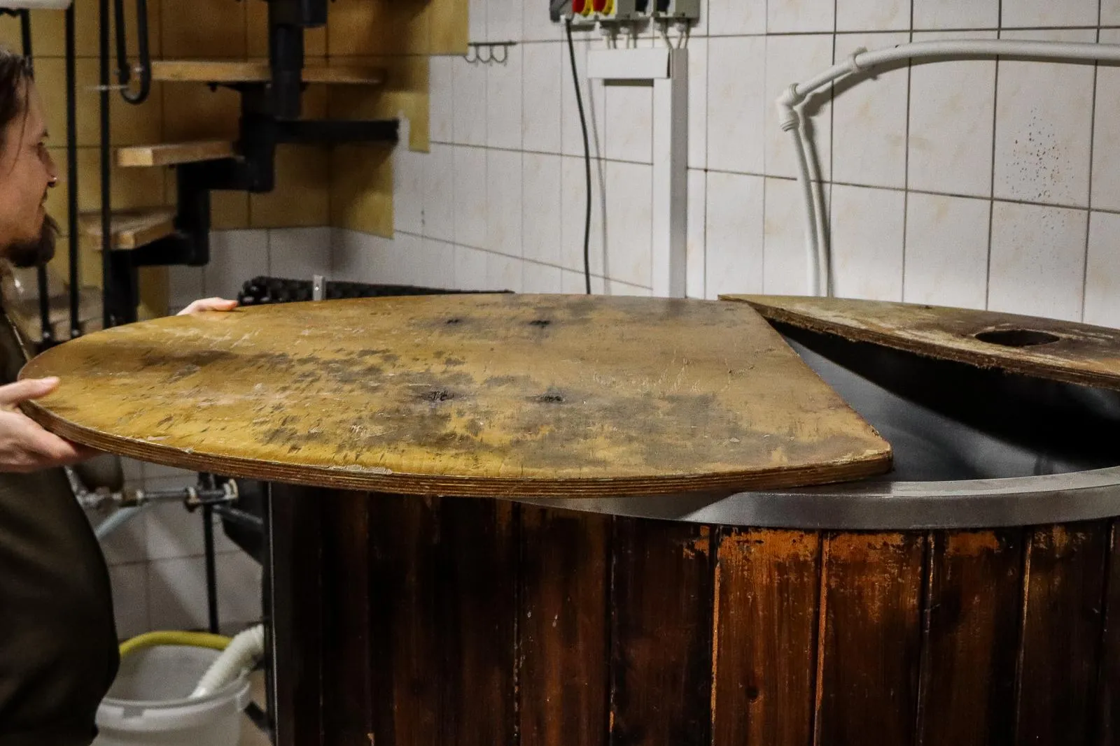 A brewer opens the wooden lid of a large brewing vat in a small brewery.
