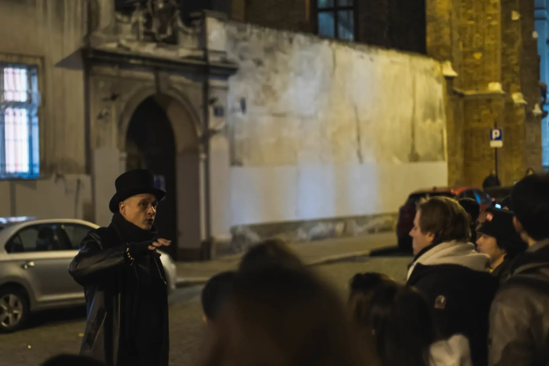 Tour guide in top hat and long coat speaking to a group at night on a cobblestone street in Krakow. Historic buildings are...
