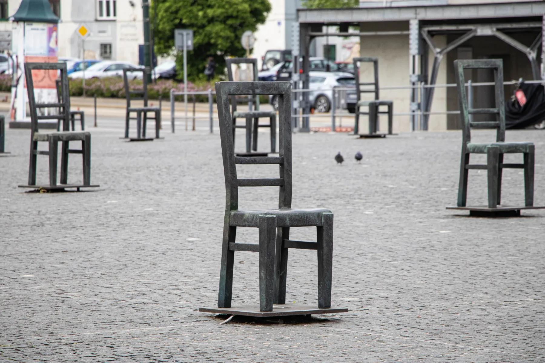 Metal chair sculptures fill a cobblestone square, with two pigeons visible and buildings in the distance.