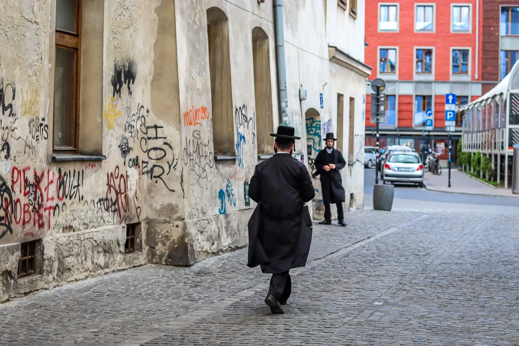 Two Orthodox Jewish men on a cobblestone street in Krakow's Kazimierz district, near a wall covered in graffiti.