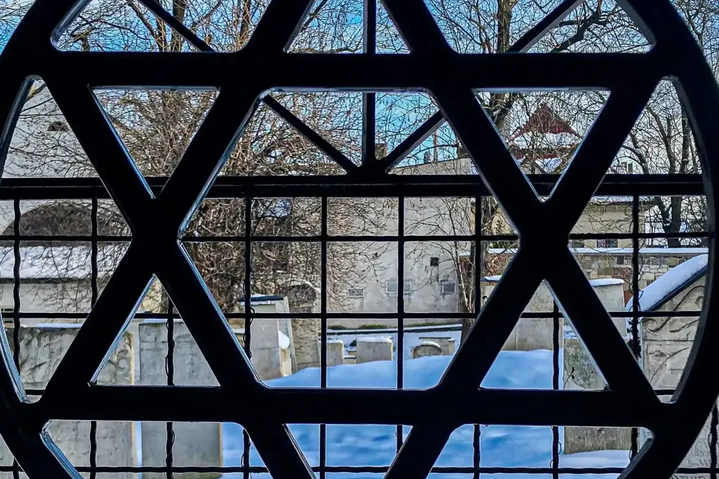 View through a window with a Star of David grille. Outside, a snow-covered cemetery with gravestones and bare trees.