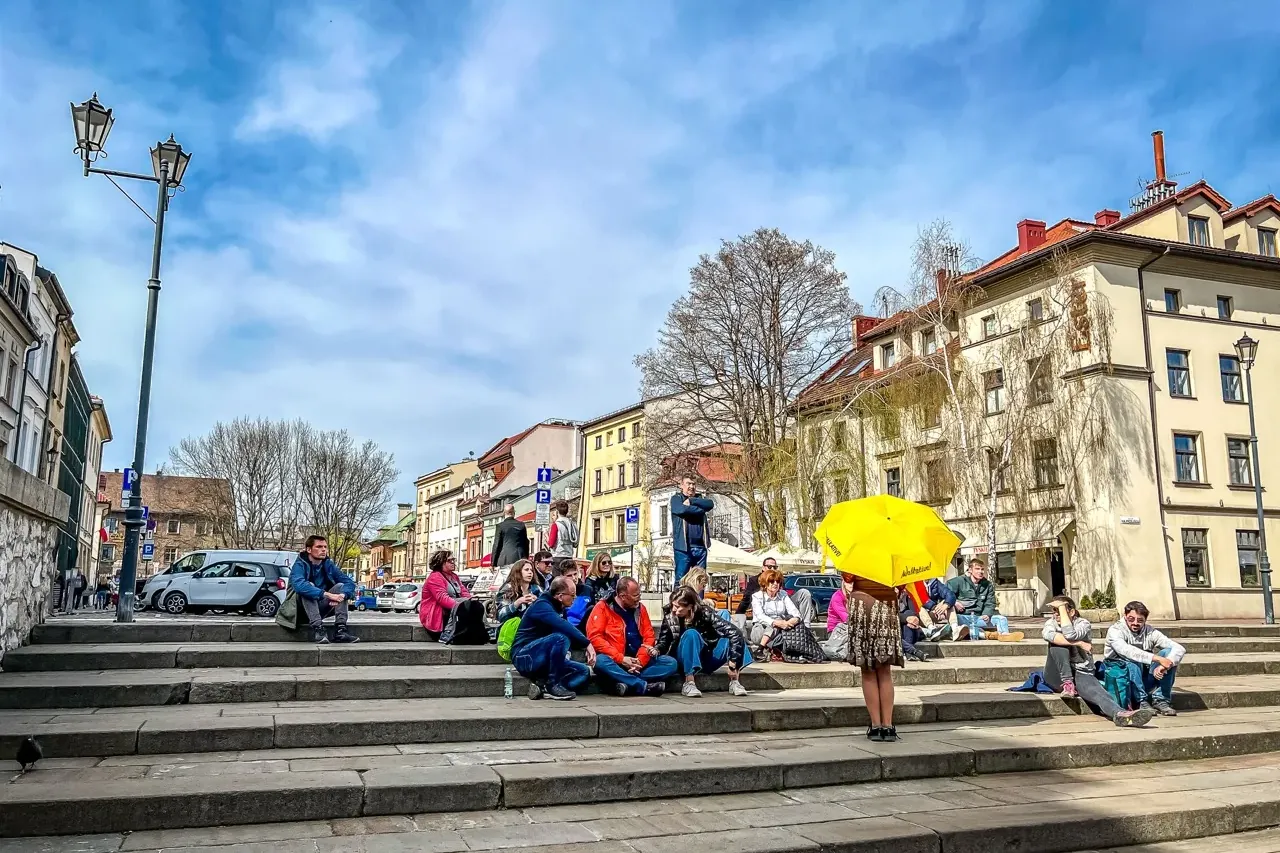 People sit on wide stone steps in a city square, historic buildings and a lamppost under a partly cloudy sky. One person h...
