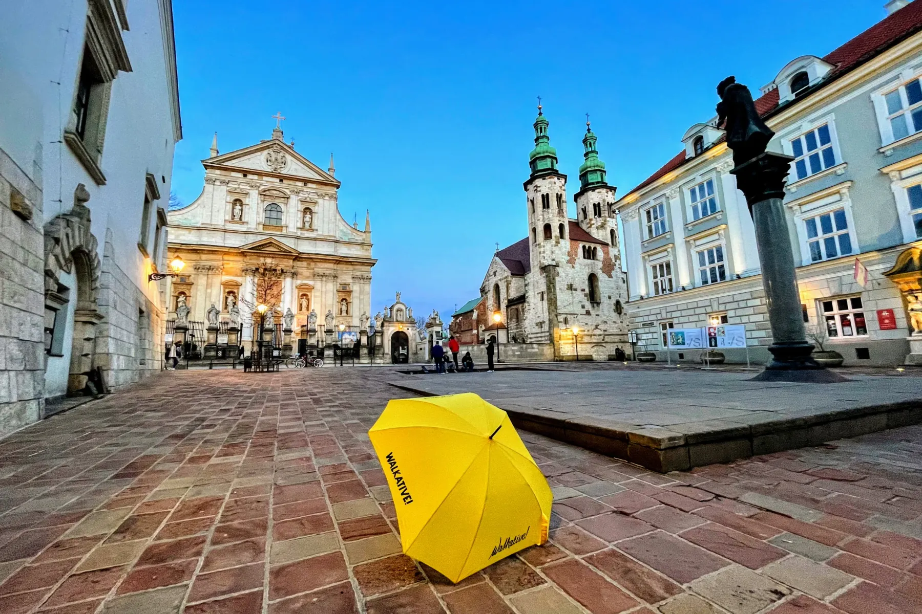 A yellow Walkative umbrella on the cobblestones of a square in Krakow, with historic churches in the background at dusk.