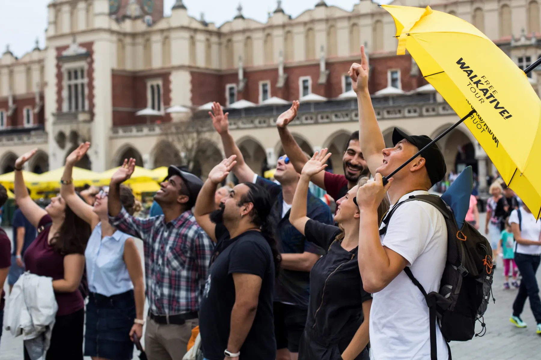 A Walkative tour guide with a yellow umbrella and a group of tourists looking up at the Cloth Hall in Krakow's Main Square.