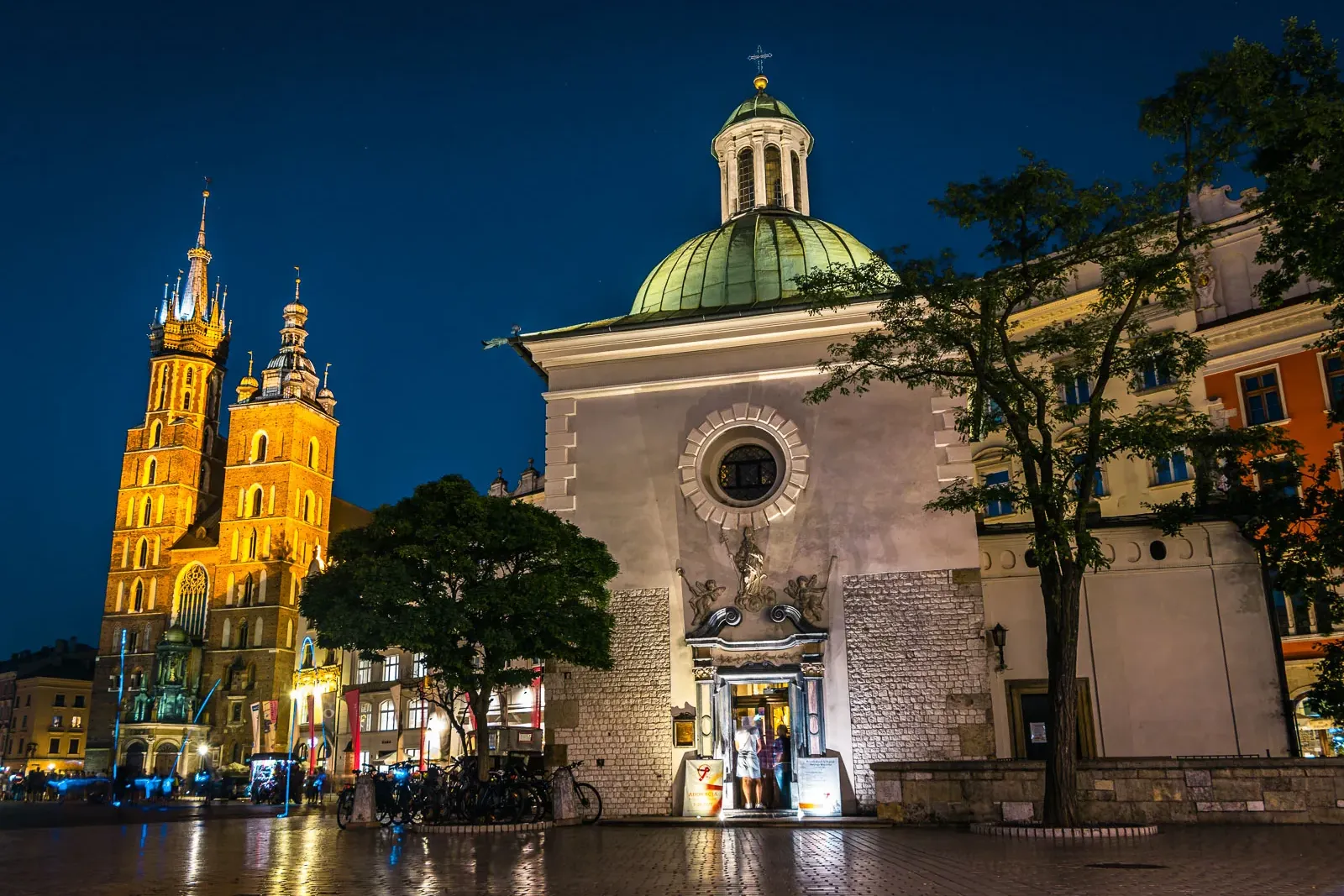 St. Mary's Basilica and St. Adalbert's Church lit up at night on Krakow's Main Market Square with wet cobblestones.