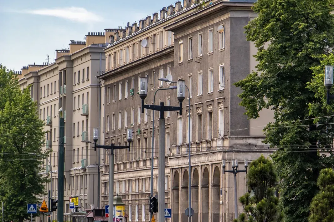 A wide street in Nowa Huta, Krakow, with long socialist realist apartment blocks, green trees, and a blue sky.