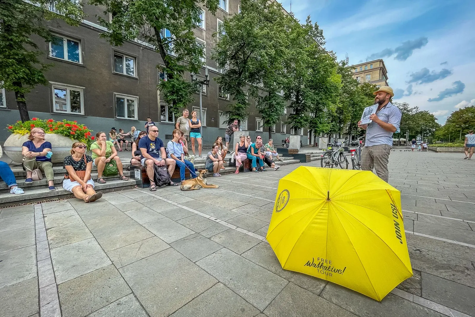 A tour guide speaks to a group of people sitting on steps in a city square, with a yellow Walkative umbrella in front.