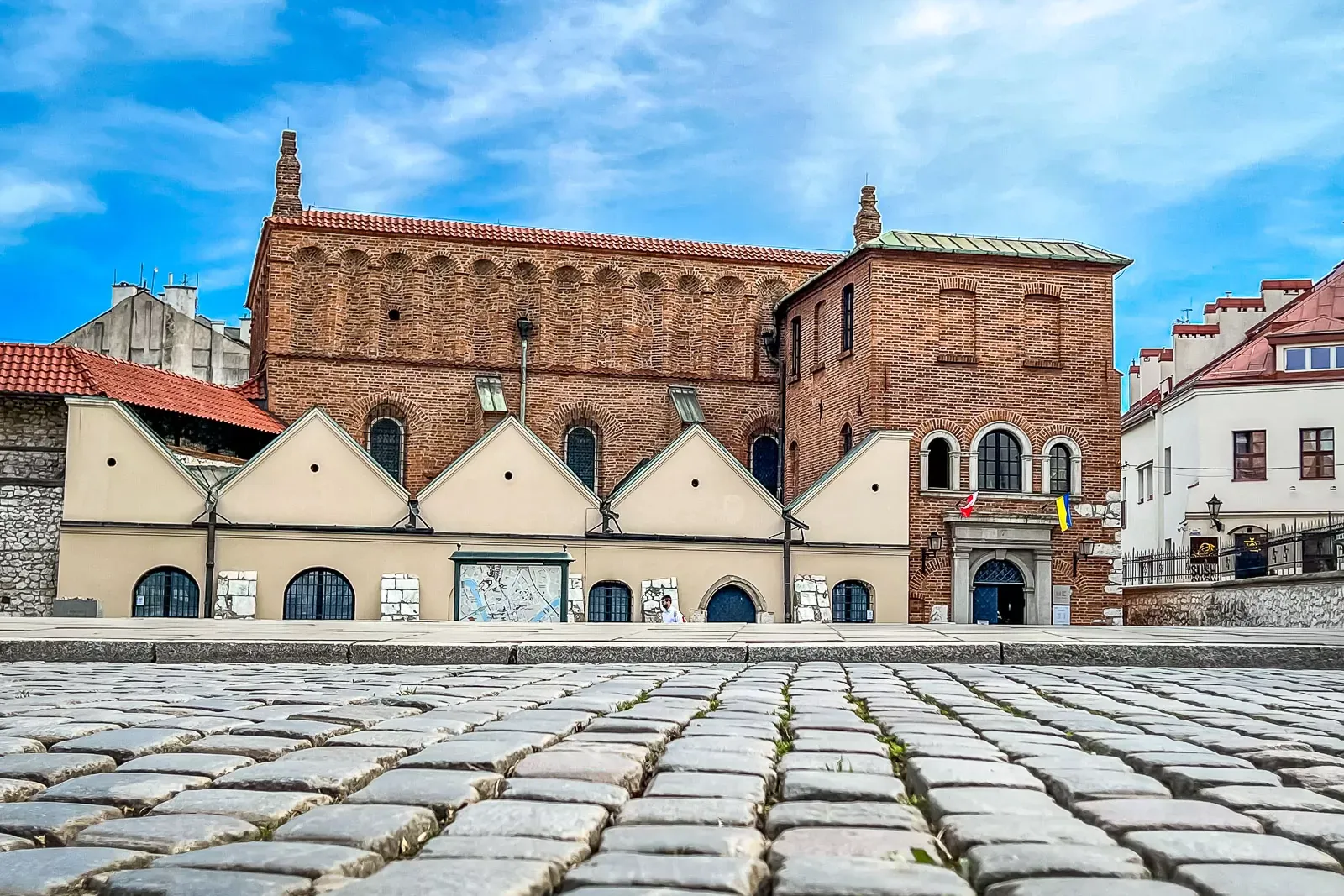 Old brick synagogue with arched windows, red roof, and cobblestone foreground under a blue sky in Krakow.