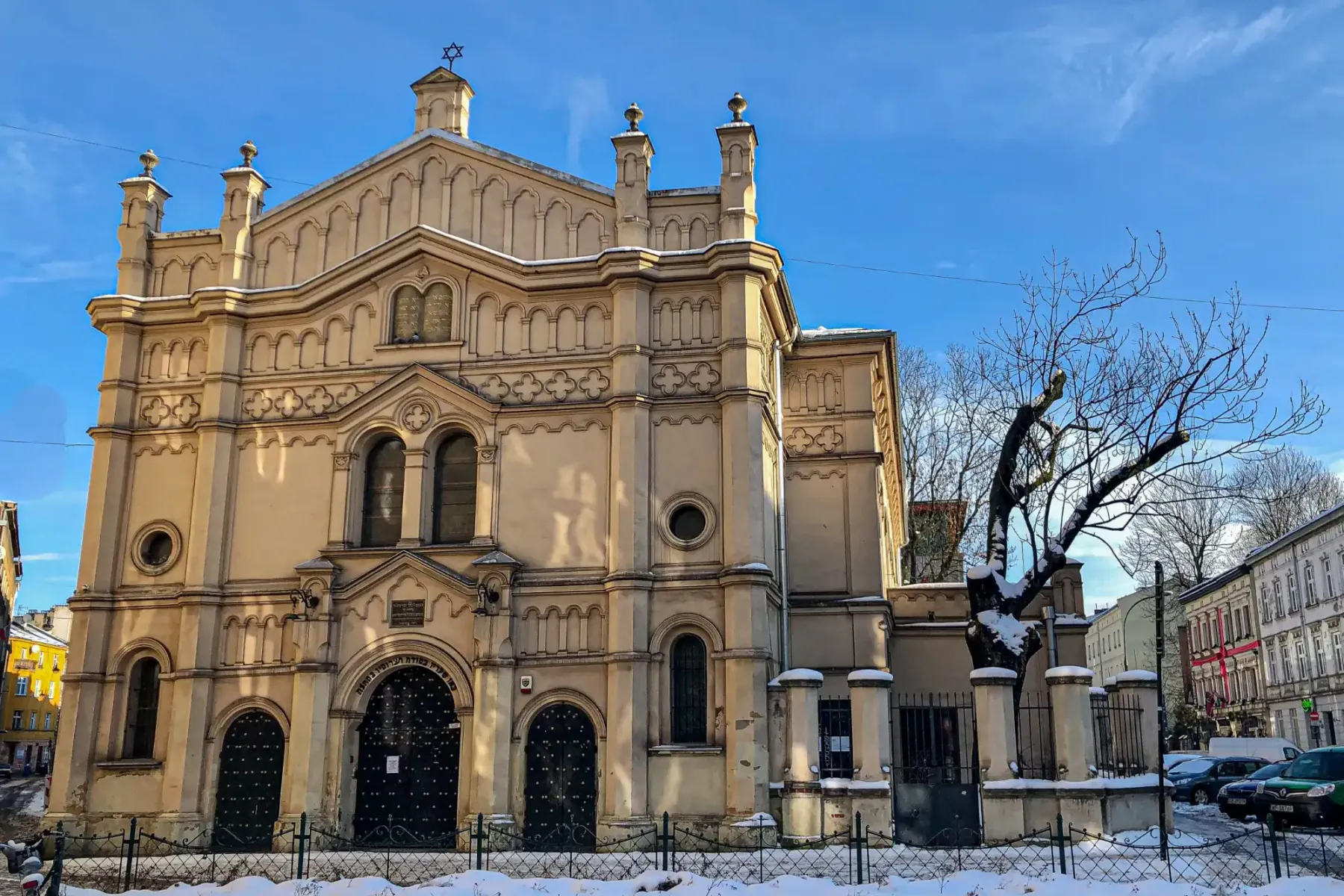 Exterior view of a large, ornate synagogue with a Star of David, covered in snow, under a clear blue sky in Krakow.