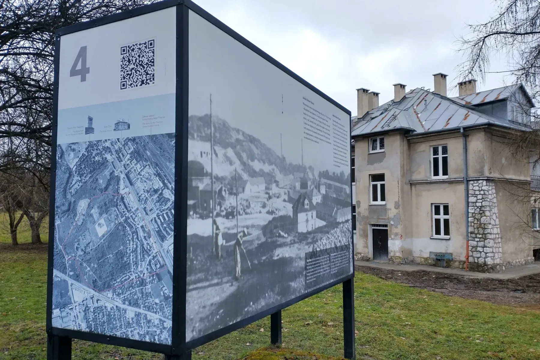 An information board with a map and historical photos stands on a lawn in front of the 'Grey House' at the Plaszow camp site.