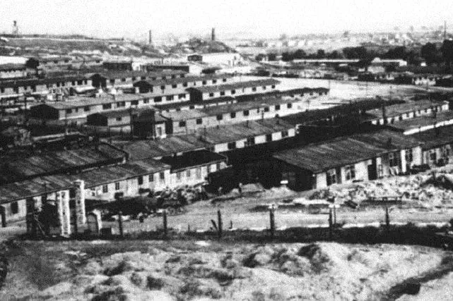 Black and white aerial view of Plaszow Concentration Camp with numerous barracks, fences, and distant industrial structures.