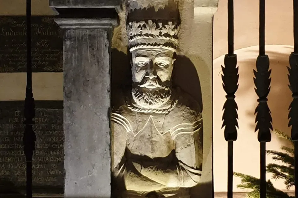 A weathered stone sculpture of a bearded king with a crown, built into a pillar and illuminated from below at night.