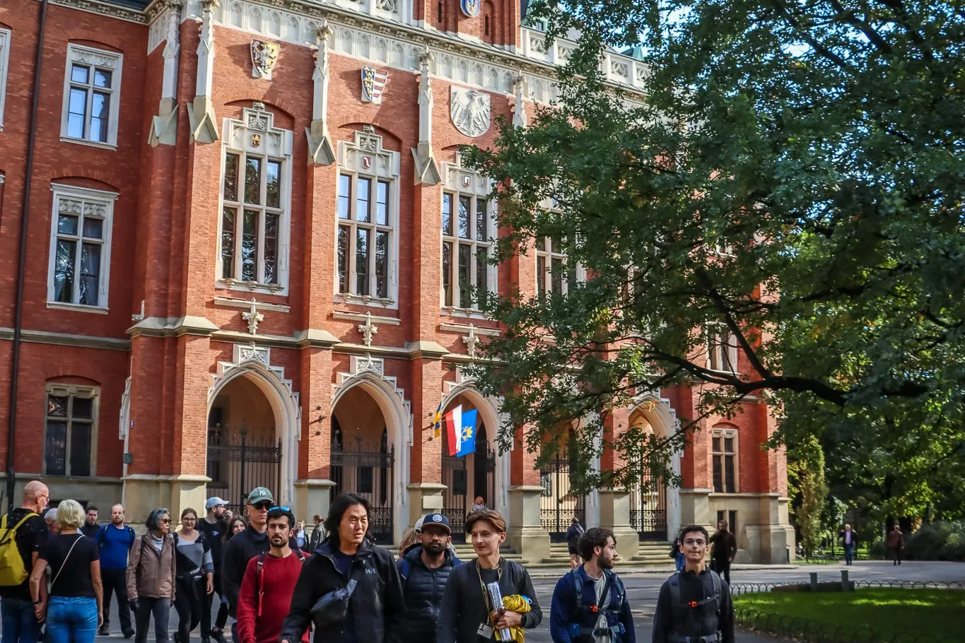 A tour group walks past the red brick neo-Gothic facade of the Collegium Novum in Krakow on a sunny day.