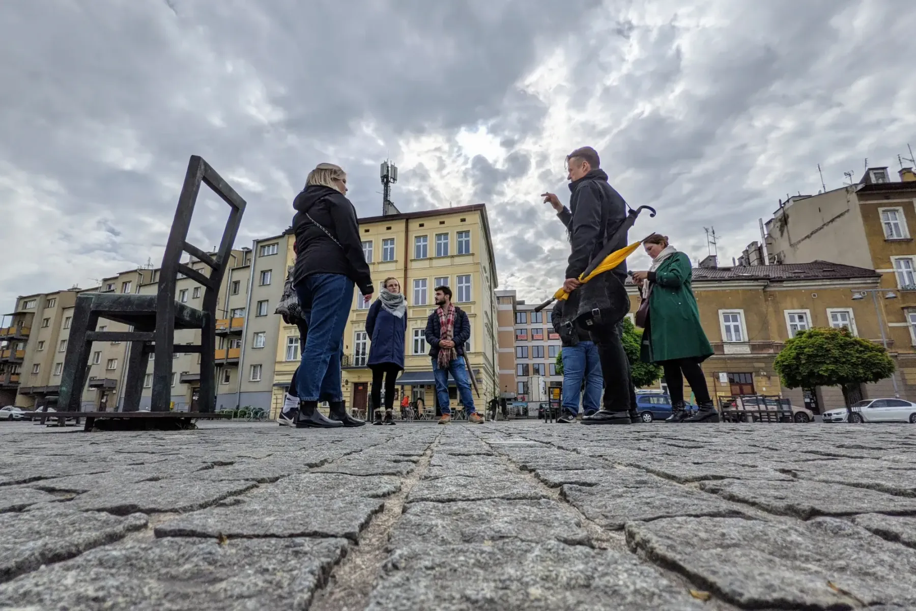 A tour guide speaks to a small group on a cobblestone square with large metal chair sculptures and buildings under a cloud...