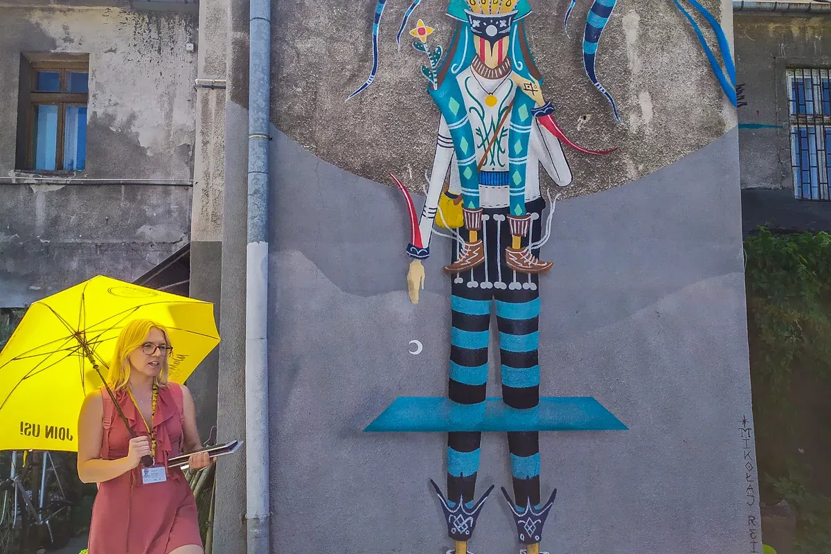 A tour guide with a yellow umbrella stands in front of a large, colorful mural on a building in Krakow.