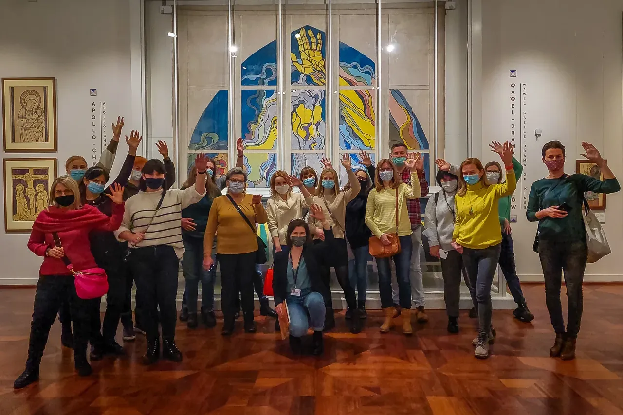 A group of tourists wearing face masks waves inside a museum in front of a large, modern artwork.