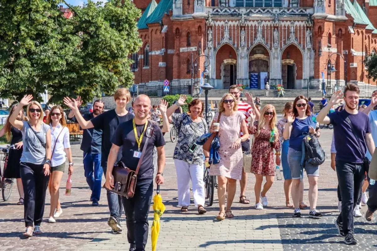 A tour guide with a yellow umbrella leads a group waving at the camera in front of a large brick church in Krakow.