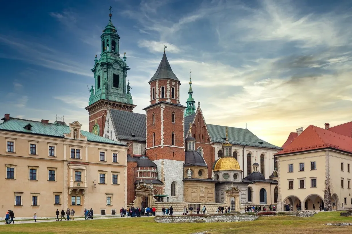 The Wawel Cathedral complex on a partly cloudy day, with its various towers and chapels seen from a grassy lawn.