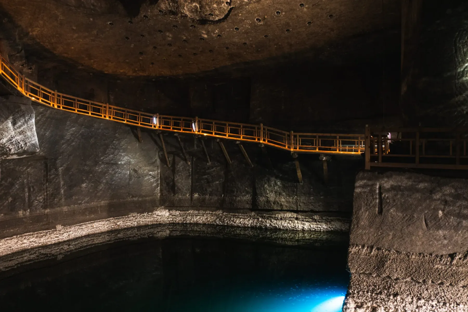 An illuminated wooden walkway runs along the cavern wall above an underground brine lake in the Wieliczka Salt Mine.