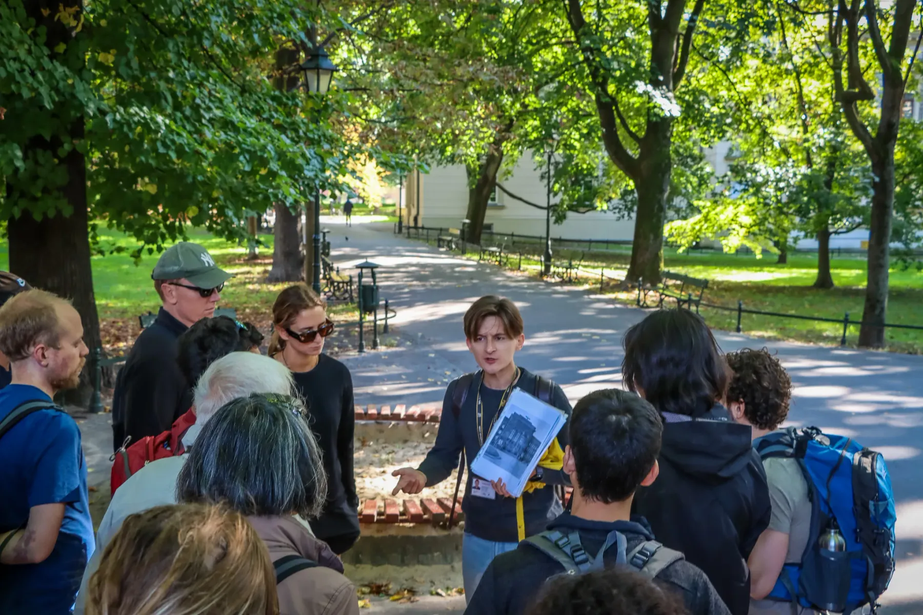 A tour guide shows historical photos to a group of people on a path in a sunlit park in Krakow.