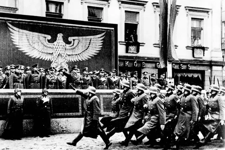 Black and white photo of a Nazi military parade on a street in Krakow. Soldiers march past a stand with officers and a lar...