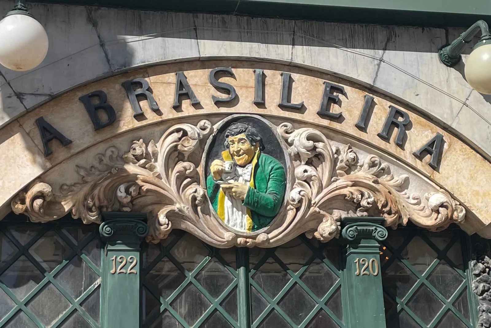 Facade of Café A Brasileira in Lisbon, showing its arched sign, a central relief of a man with a cup, and green-framed win...