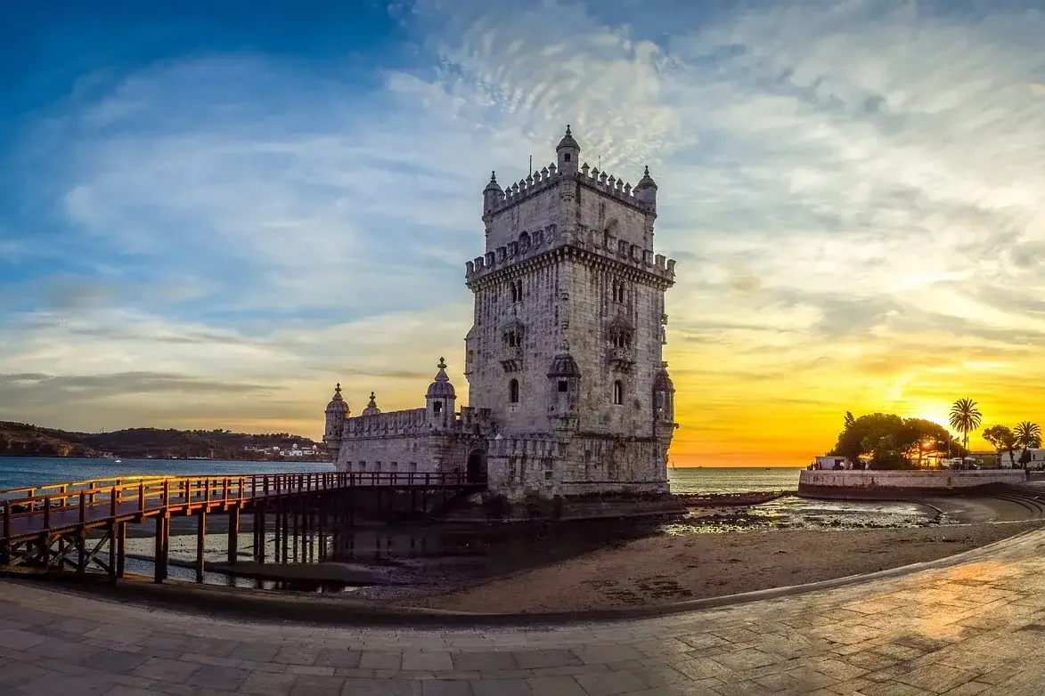 Historic Belém Tower in Lisbon at sunset, with a wooden walkway over the Tagus river and orange-blue sky.