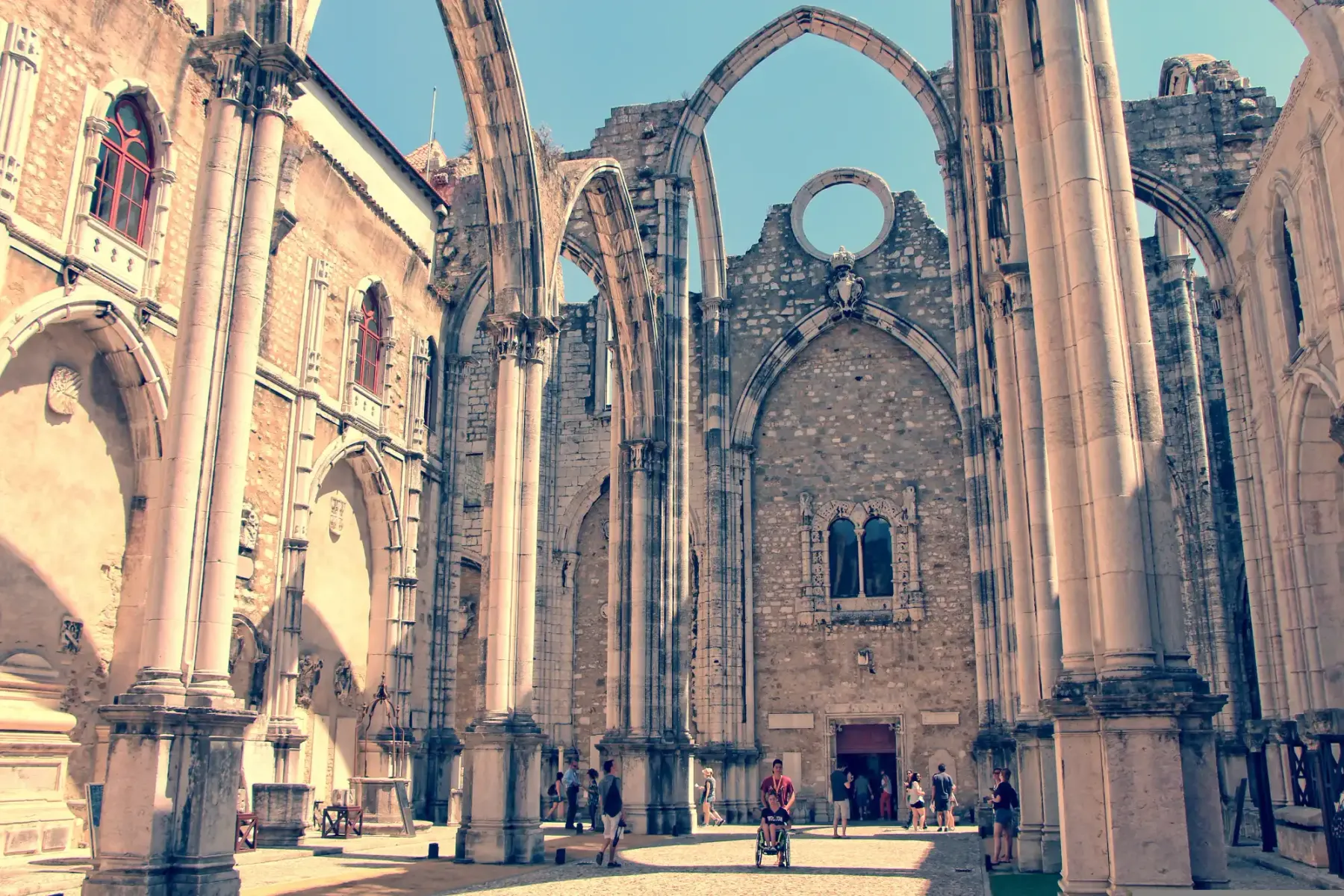 The roofless ruins of the Gothic Carmo Convent in Lisbon, with stone arches reaching against a clear blue sky.