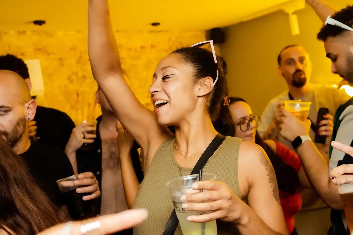 Group of people dancing and holding drinks in a warmly lit bar. A woman in the foreground smiles with one arm raised.