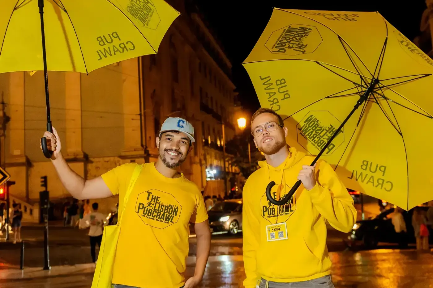 Two men in yellow 'The Lisbon Pubcrawl' apparel holding yellow umbrellas, standing on a wet street at night in Lisbon.