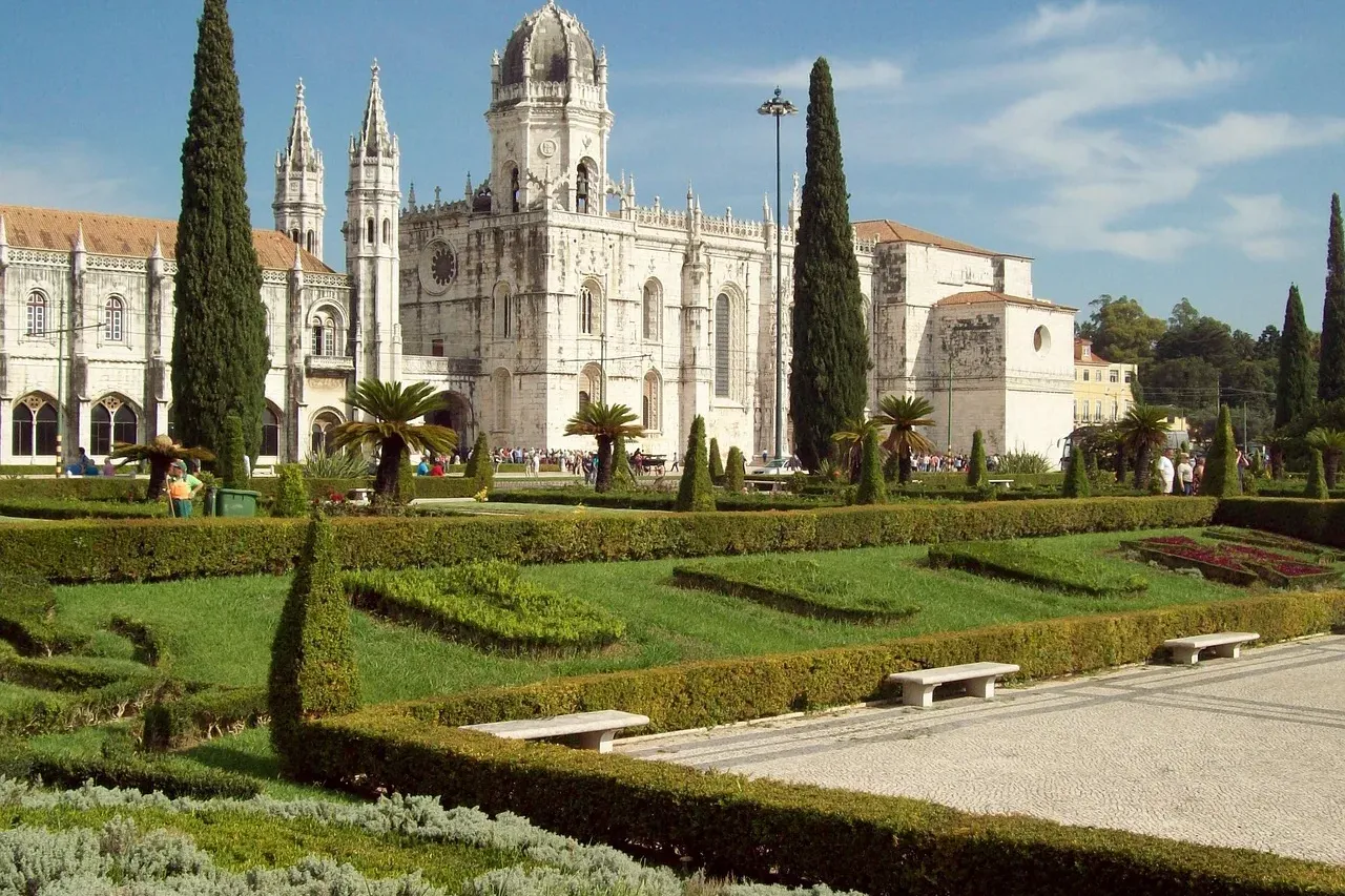 Jerónimos Monastery in Lisbon with manicured green gardens, cypress trees, and visitors on a sunny day.