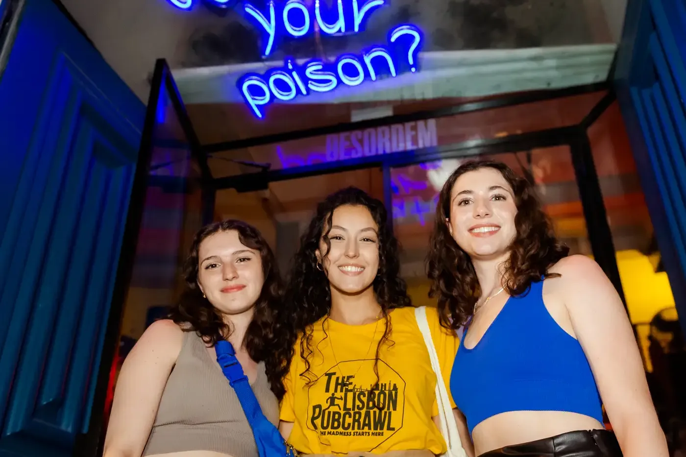 Three young women smiling in front of a blue neon sign that reads 'What's your poison?' above a blue door frame.