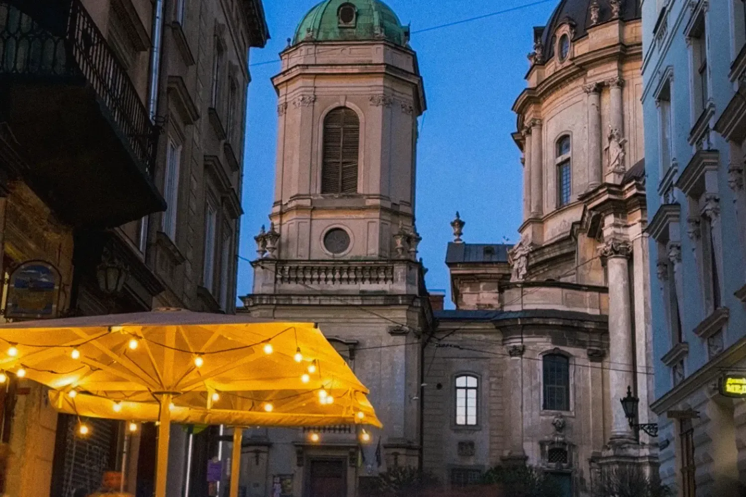 A busy street cafe in Lviv at dusk, with the Dominican Cathedral in the background under a deep blue sky.