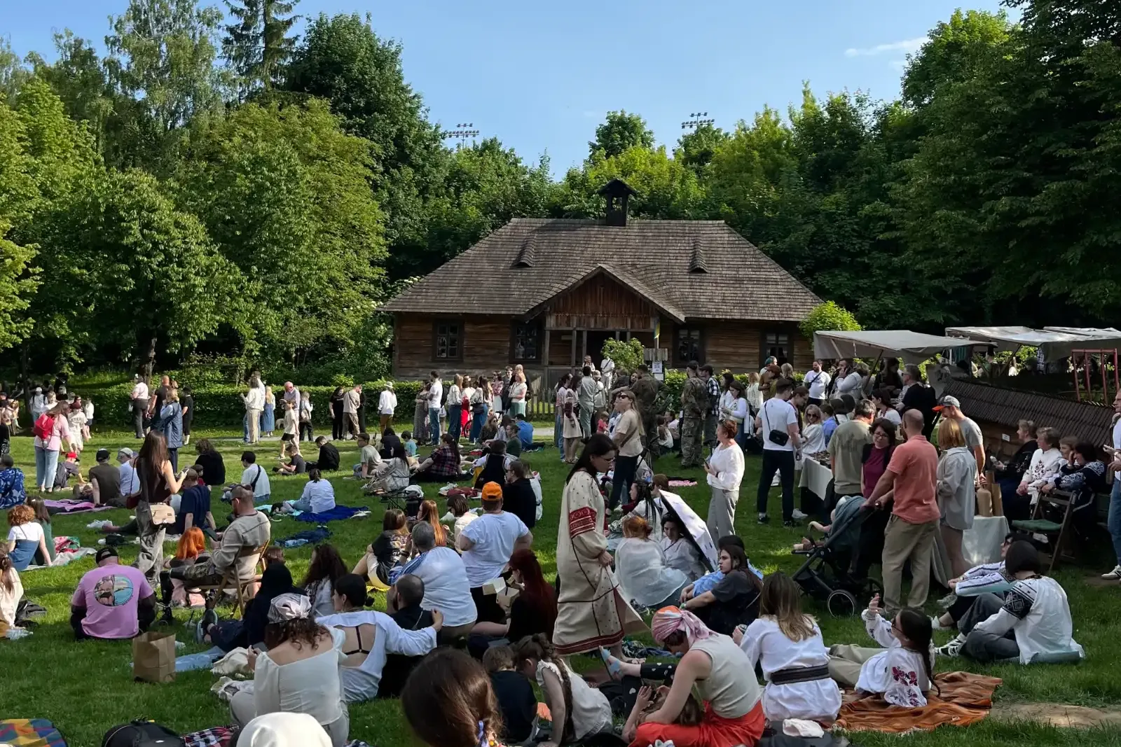 A large crowd of people gathered on a grassy lawn in front of a traditional wooden house on a sunny day.