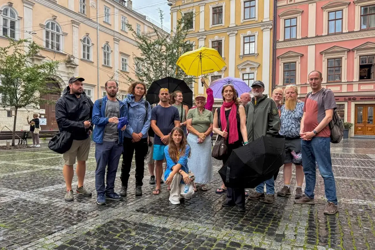 A group of about 15 adults stands on a wet cobblestone square in Lviv. Several hold umbrellas, with colorful old buildings...