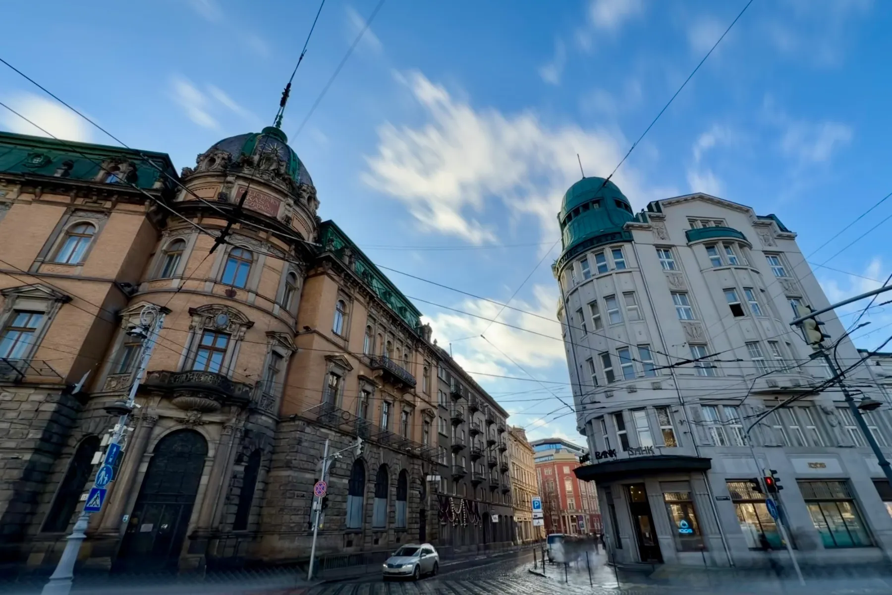 Wide-angle view of two historic buildings on a wet cobblestone street in Lviv under a blue sky with streaked clouds.