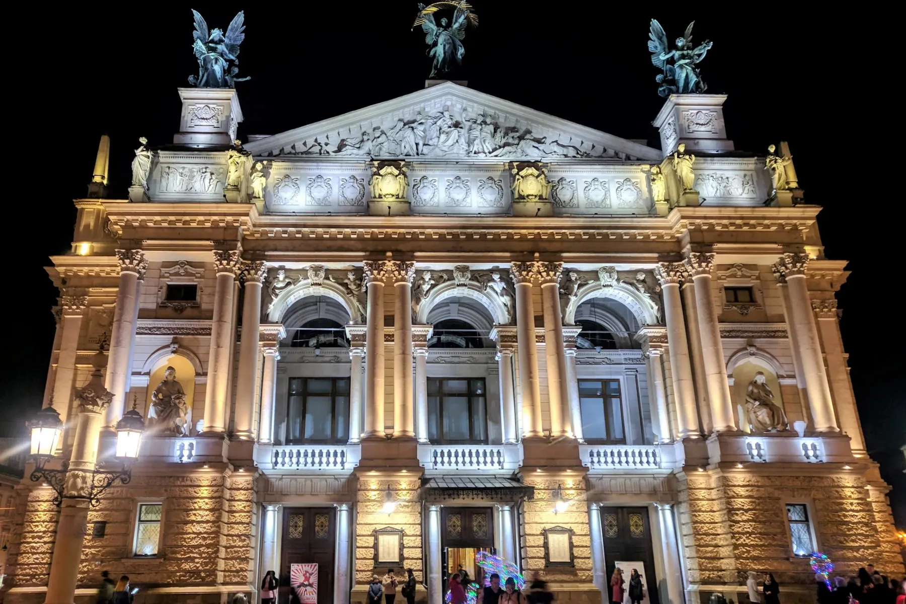 The brightly illuminated Lviv Opera House at night, with detailed sculptures and people walking in front.