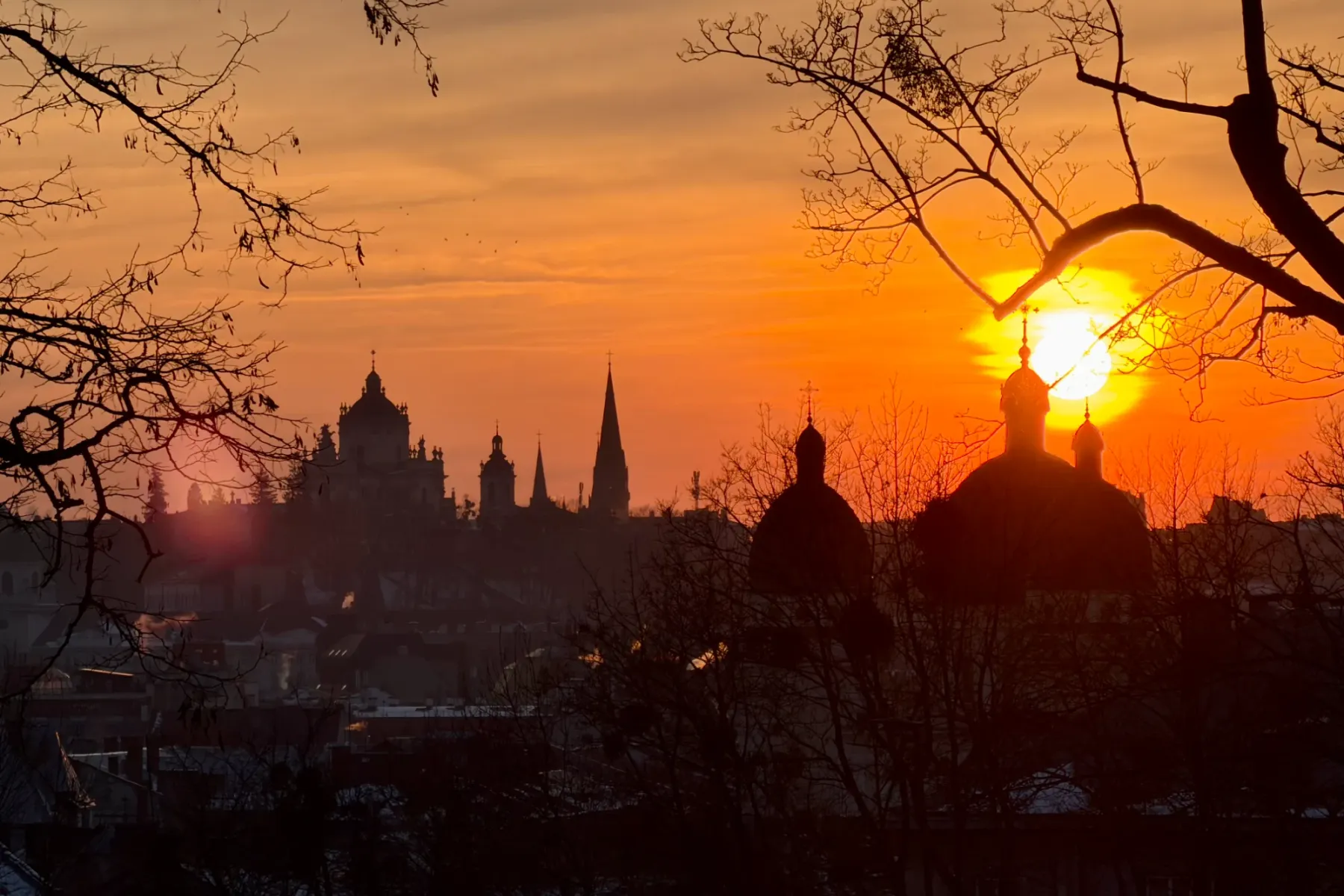 Silhouettes of church domes and spires against a bright orange sunset sky, framed by bare tree branches in Lviv.