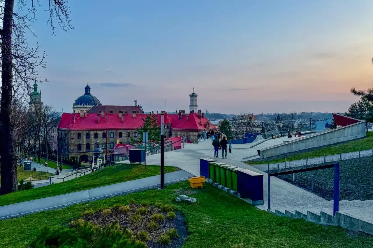 View from High Castle Hill in Lviv at sunset, showing red-roofed buildings, a dome, a tower, and modern park paths with pe...