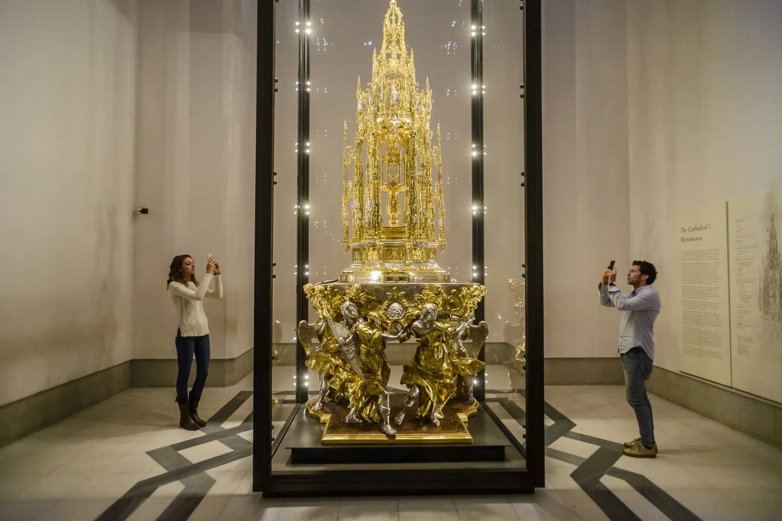 An ornate, gilded monstrance in a glass display case, being photographed by two visitors on their phones.