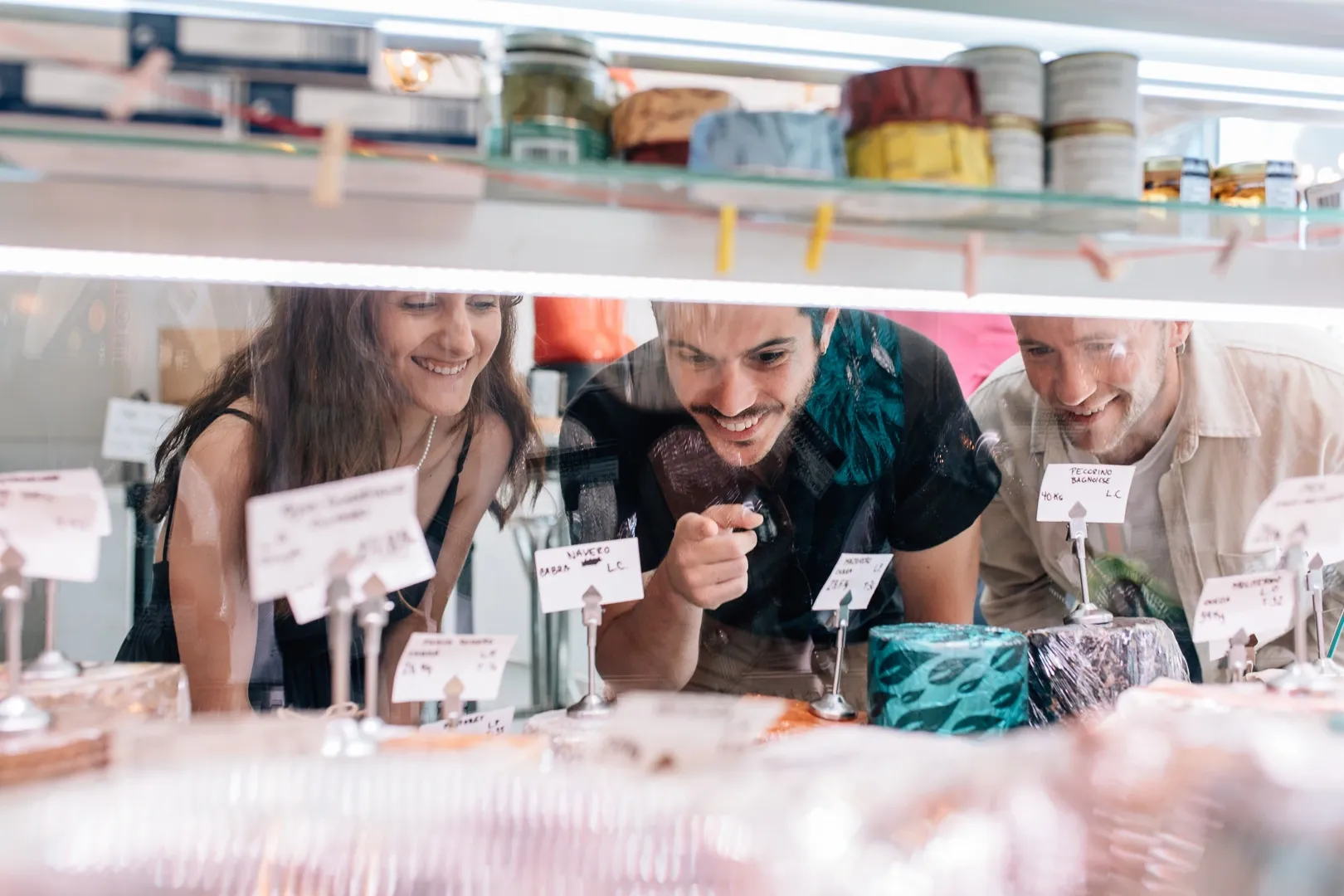 Three young people smile as they look through a glass counter at a selection of cheeses in a shop.