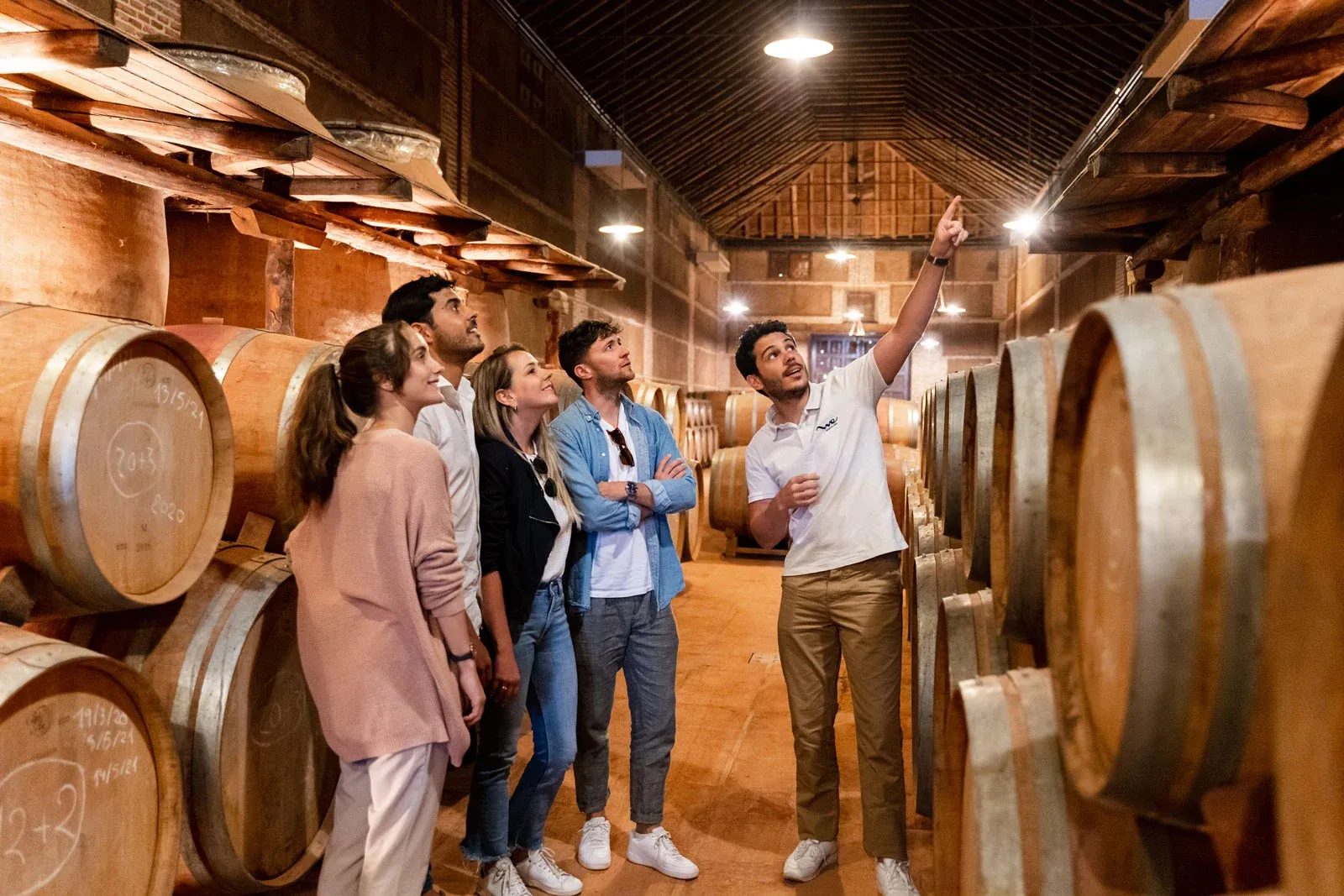A tour guide explains something to a group of visitors in a wine cellar surrounded by large wooden barrels.