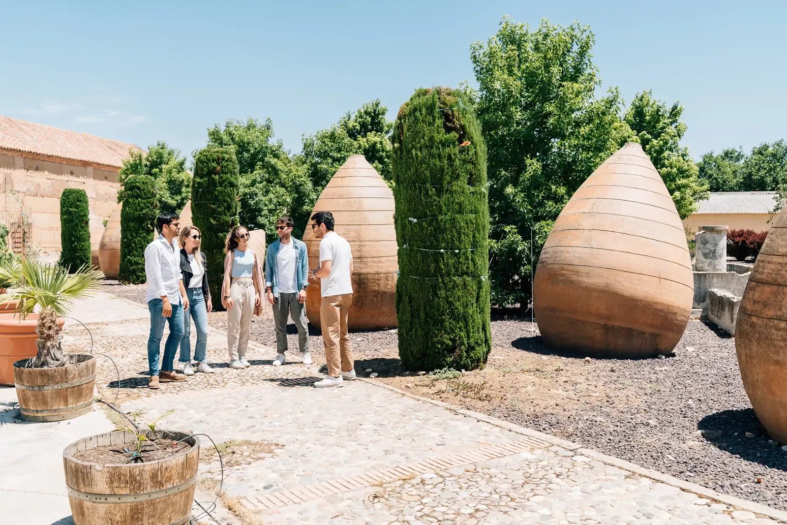 A tour guide speaks to a small group of people in a sunny courtyard with large, ancient-style clay jars.