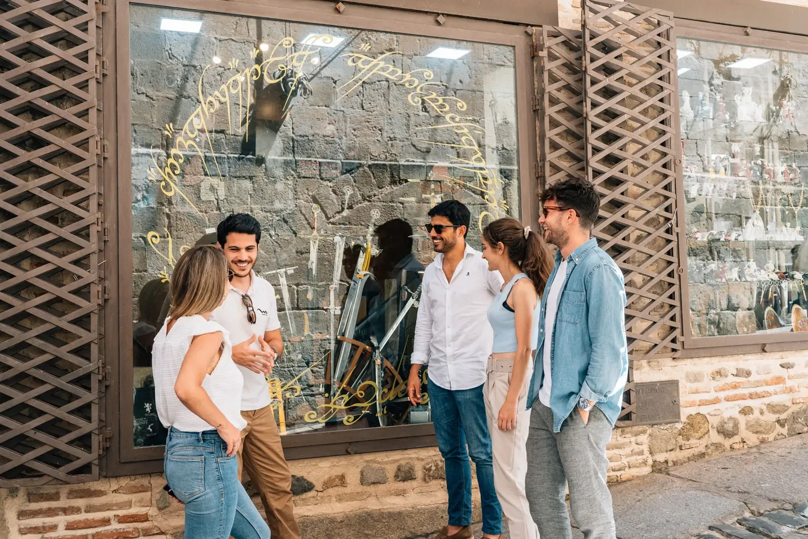 A group of five young adults talking in front of a shop with gold script on its display window.