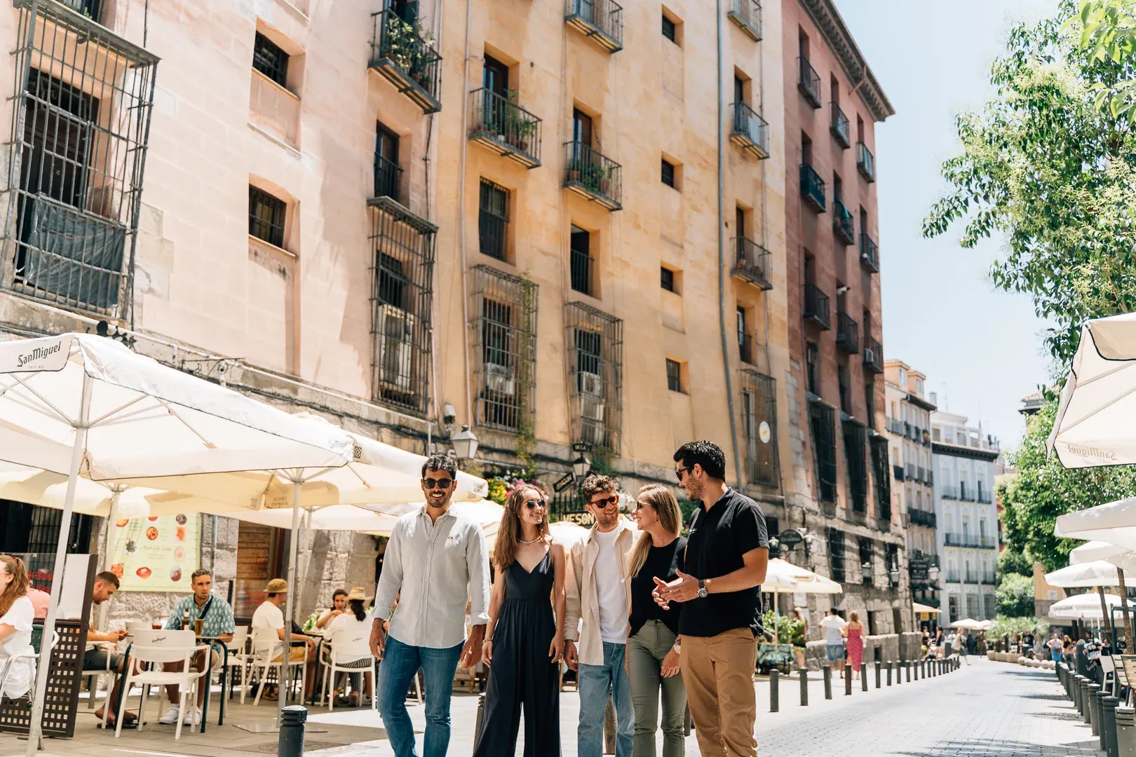 A group of five young people walk down a sunny Madrid street lined with cafe tables and historic buildings.