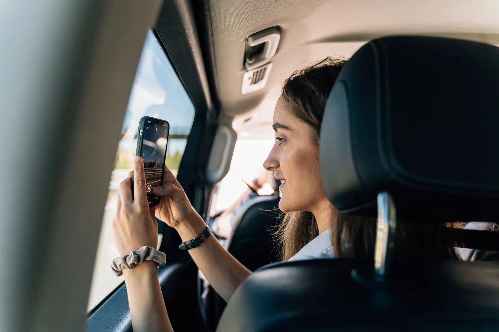 A young woman sits in the back of a car and takes a photo of the view with her smartphone.