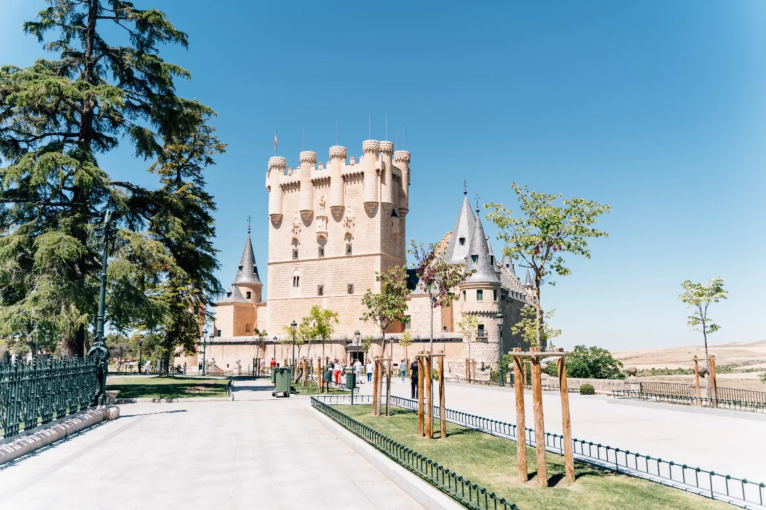 The Alcázar of Segovia, a stone castle with turrets, seen from a park on a sunny day.