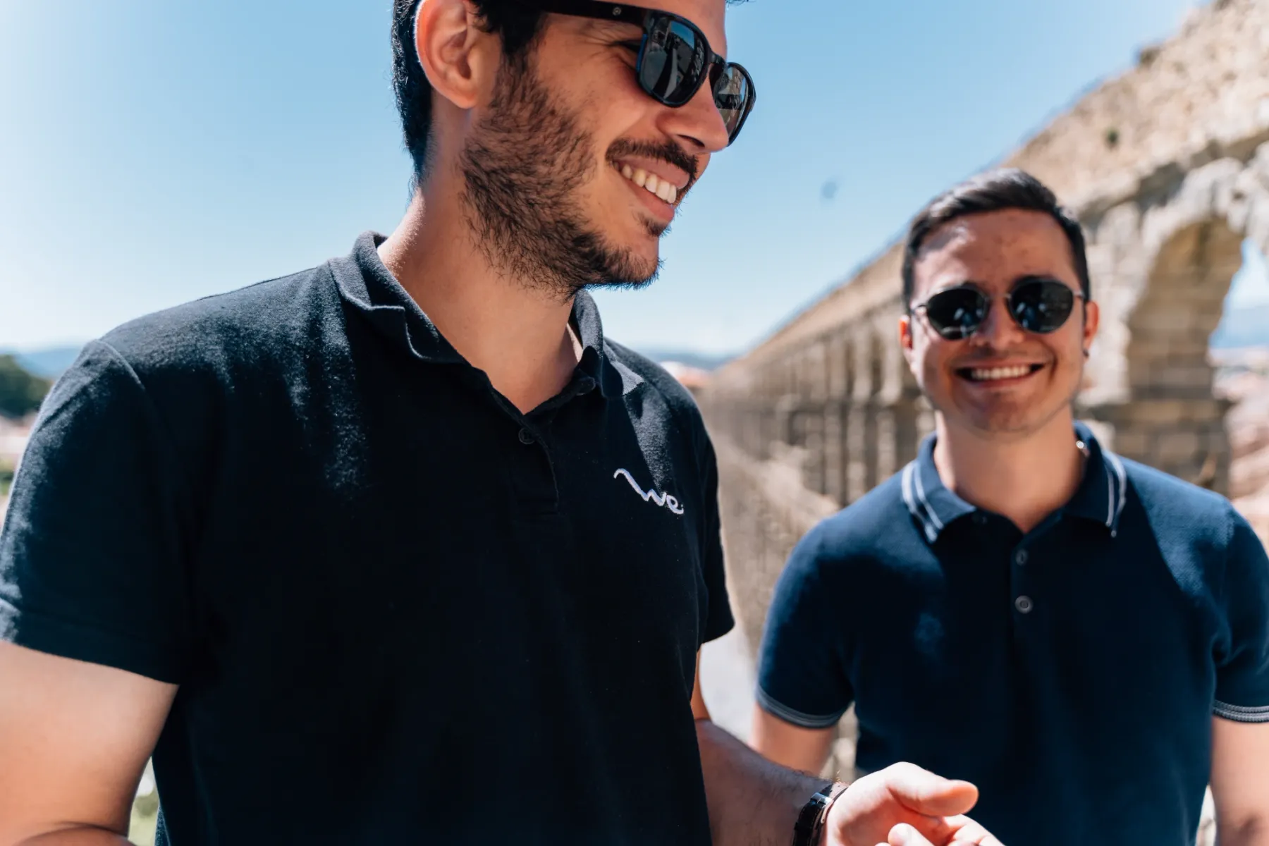 Two young men in sunglasses stand smiling in front of the Aqueduct of Segovia on a sunny day.
