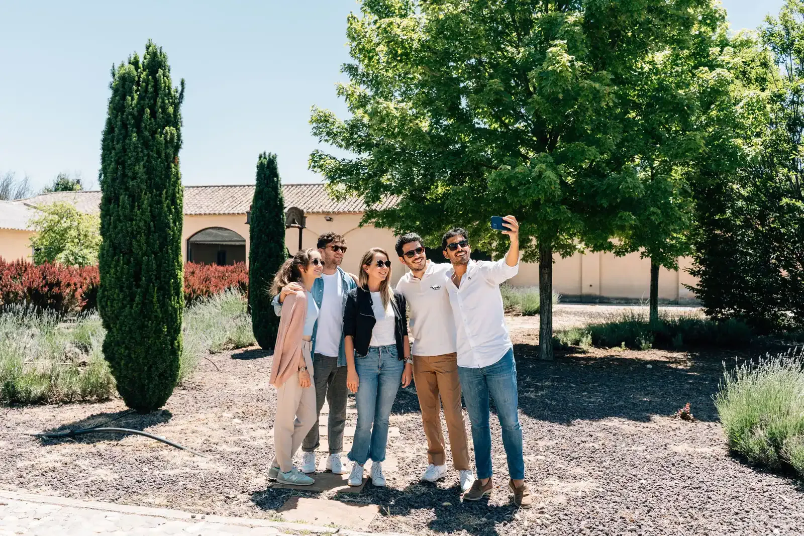 A group of five friends takes a smiling selfie in a sunny garden with cypress trees and a villa in the background.