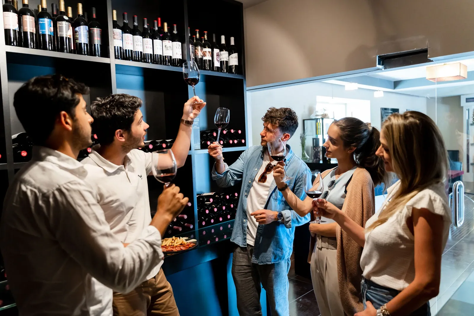 A group of people tasting red wine in a shop. One man holds his glass up to inspect the color against a wall of bottles.